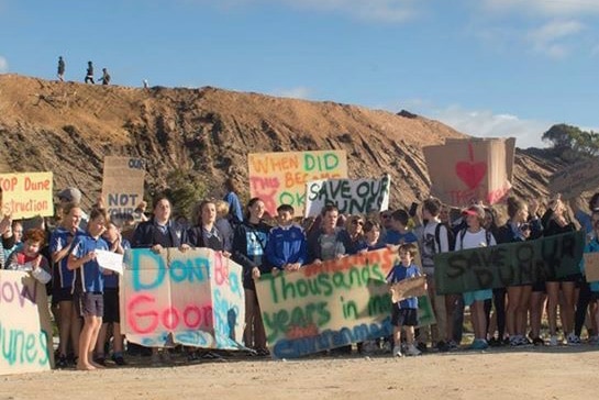 Protesters with signs standing in front of sand dunes in Golden Bay under threat from a housing development