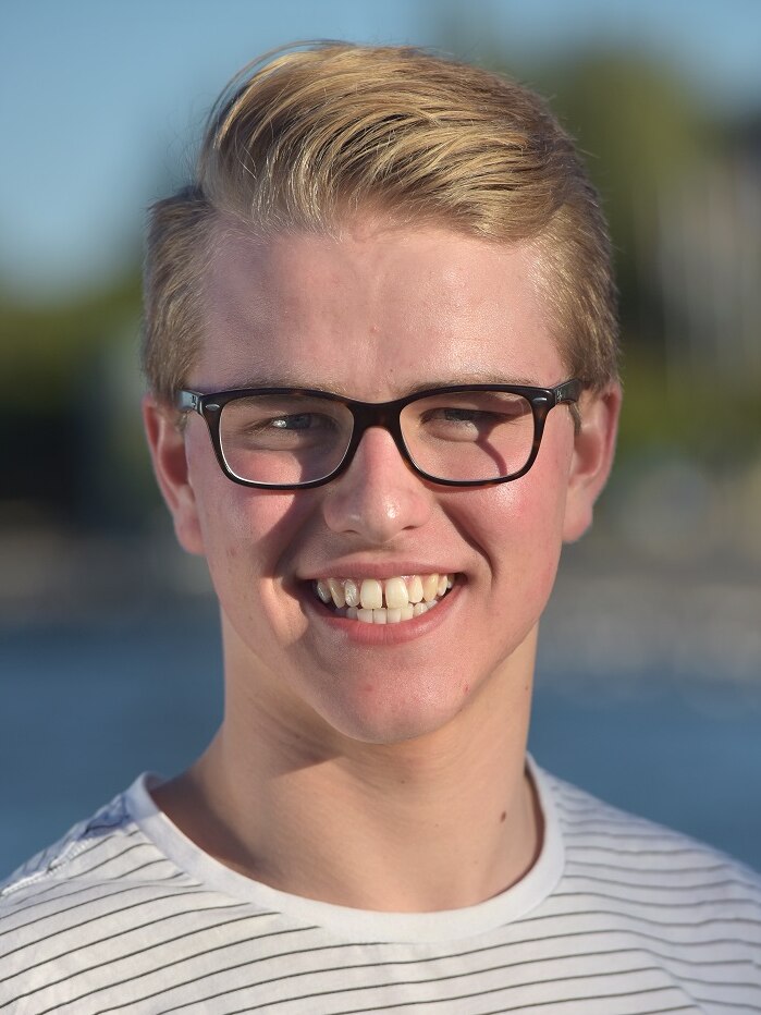 A young man with blonde hair, black glasses and a stipe shirt is smiling.