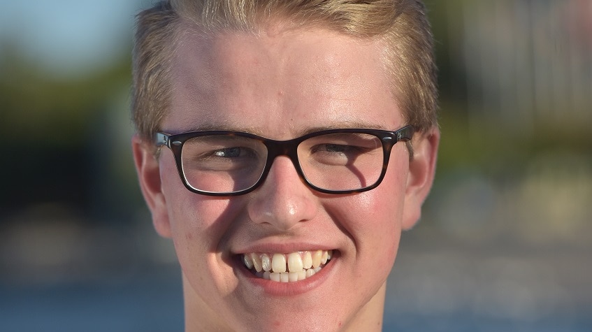 A young man with blonde hair, black glasses and a stipe shirt is smiling.