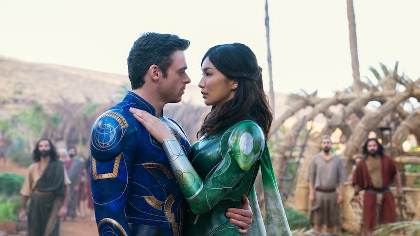 Richard Madden and Gemma Chan, wearing blue and green superhero costumes respectively, are in an intimate embrace