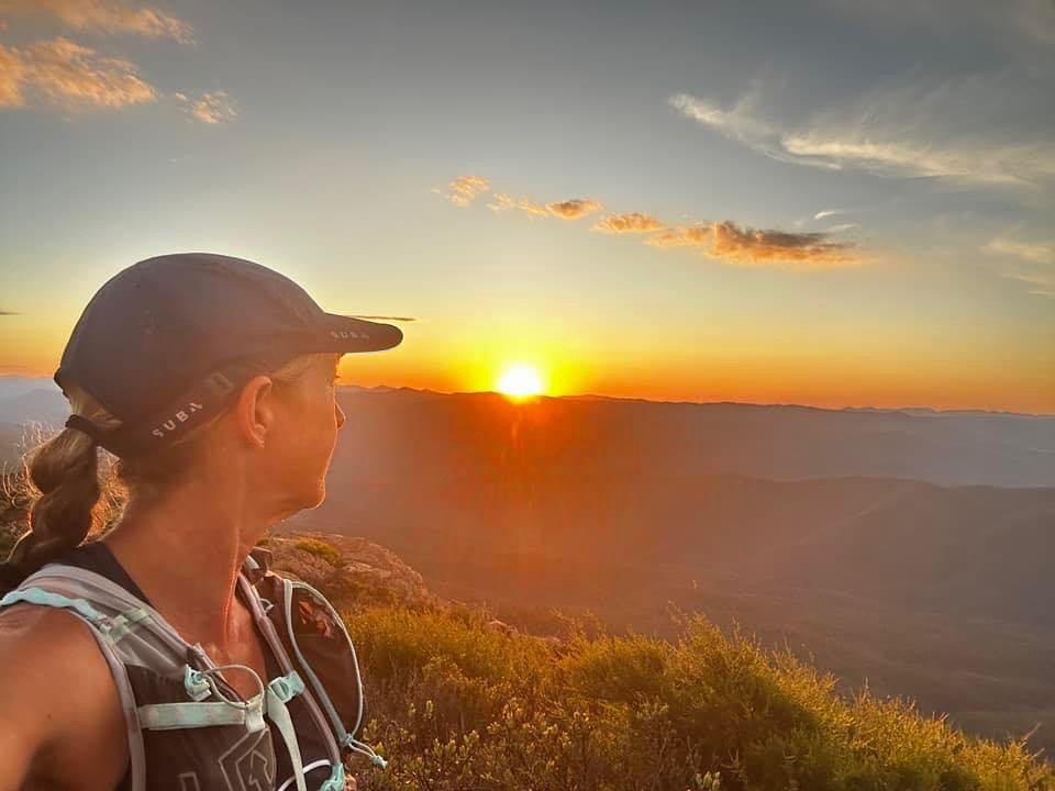 Shiree smiles, looking at a sunset over the bush, wearing a hat.