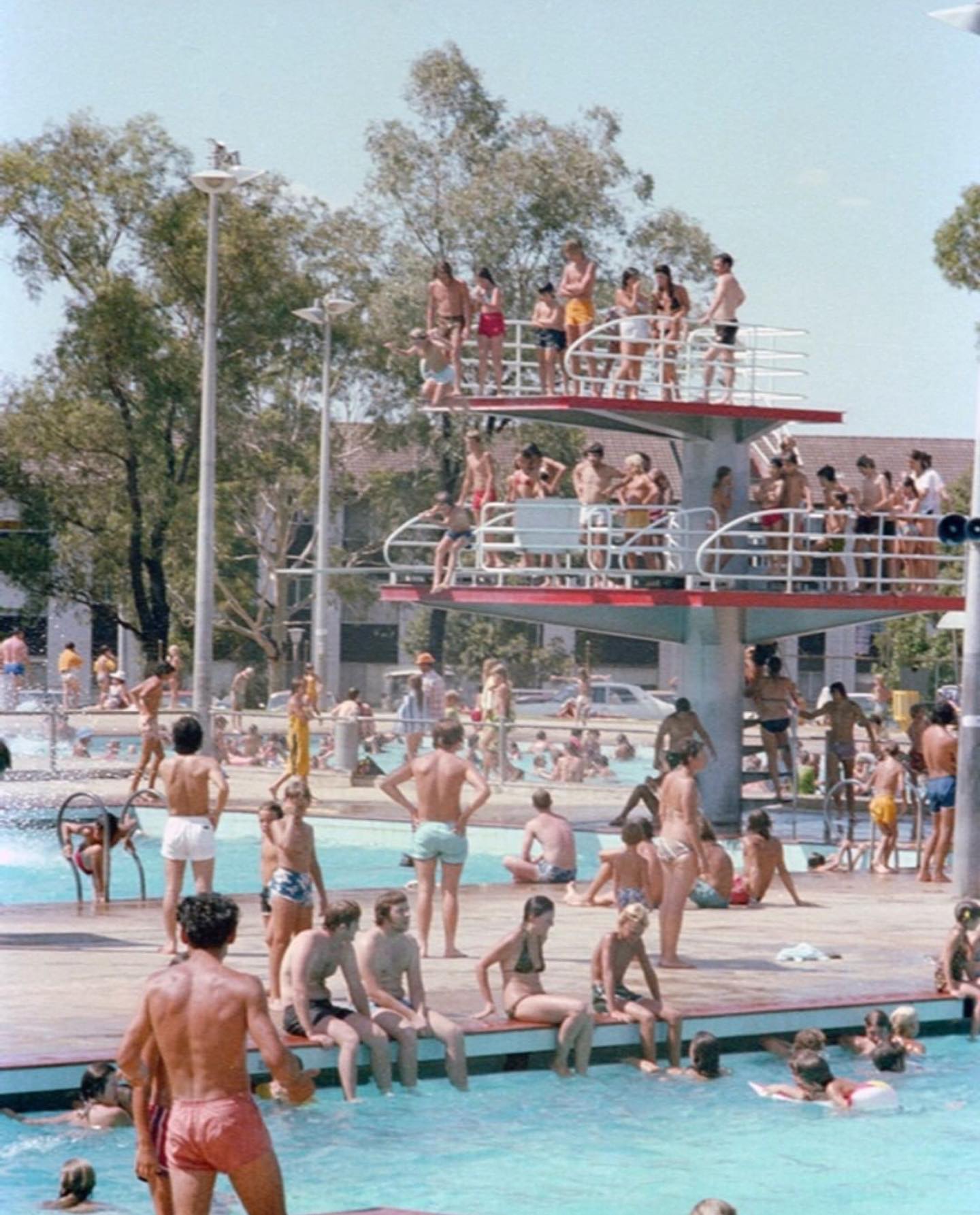 A 1979 photo of a two-tier diving platform packed with people in board shorts and bikinis. 