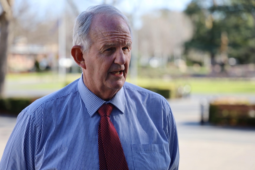 A white-haired man in a blue and white checked shirt, red tie being interviewed