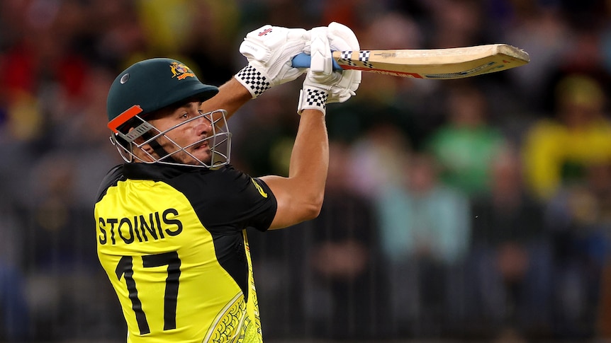 A man in a green and yellow Australian T20 cricket uniform lifts his bat high over his shoulder during a shot.