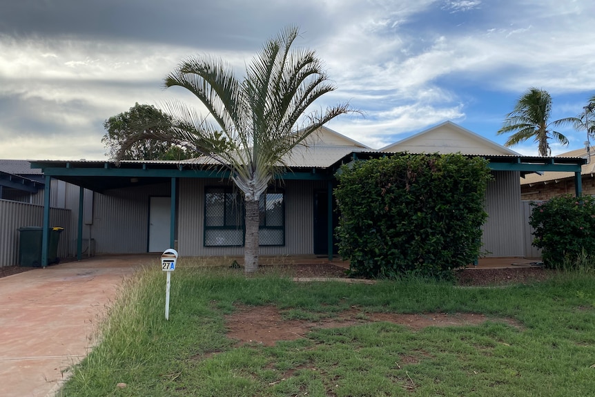 A grey tin house with green window trims and poles with a palm tree in the front yard and patchy grass and red dirt.