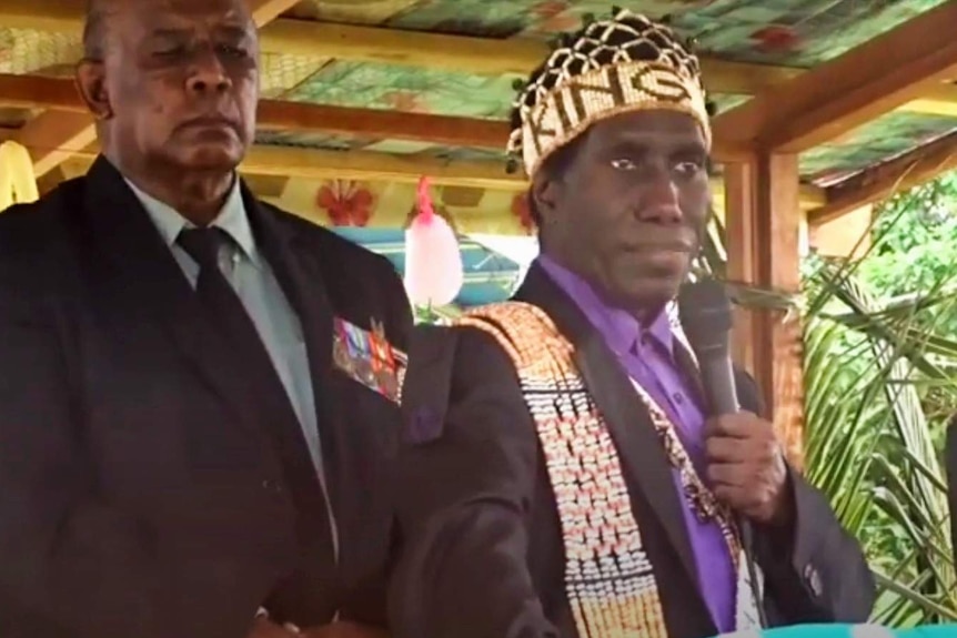 Conman Noah Musingku, wearing a crown that appears to be woven, speaks into a microphone at a podium. He wears a suit.