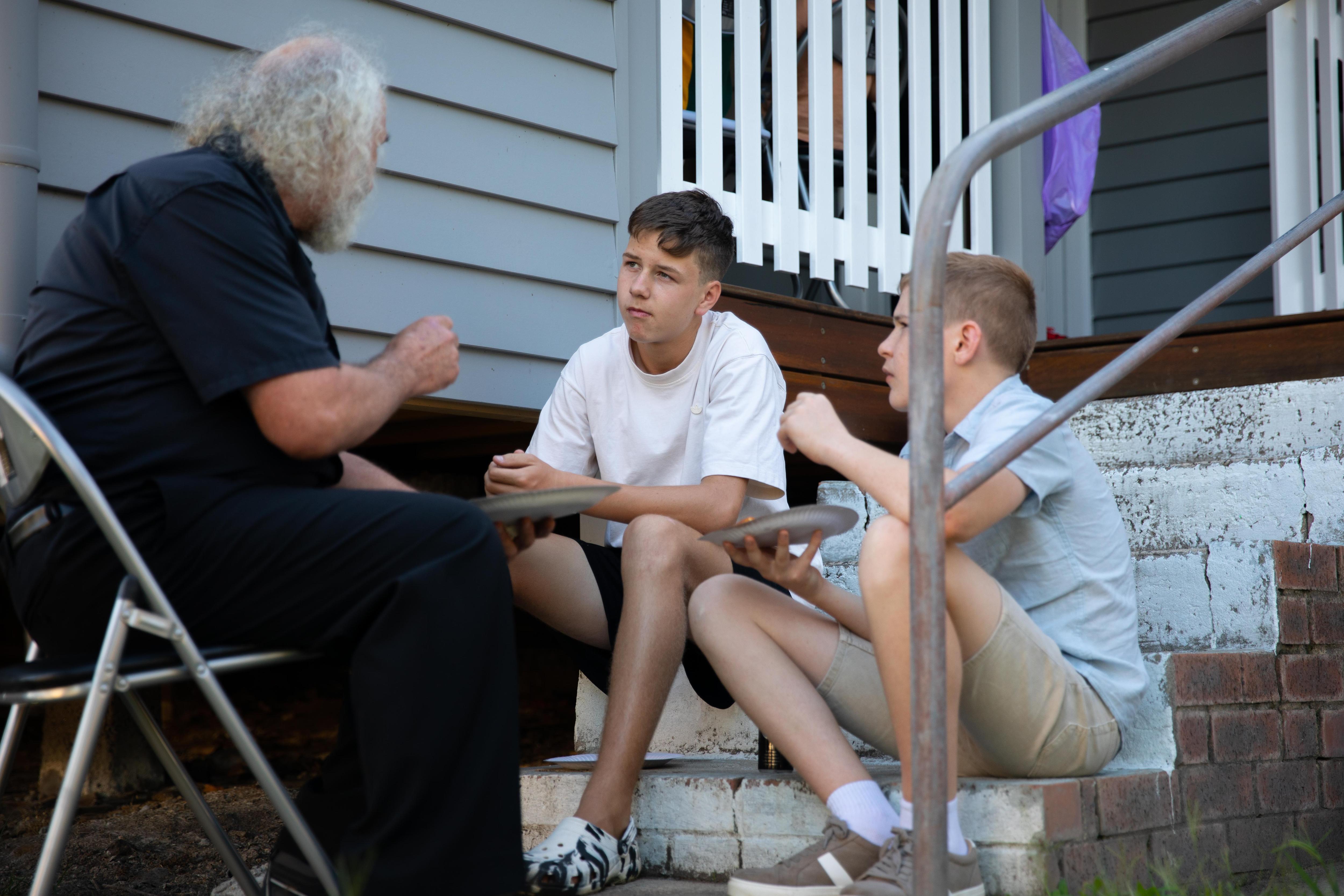 Two male teens sit on steps eating pizza while chatting with professor Wayne Burton who sits on a chair 