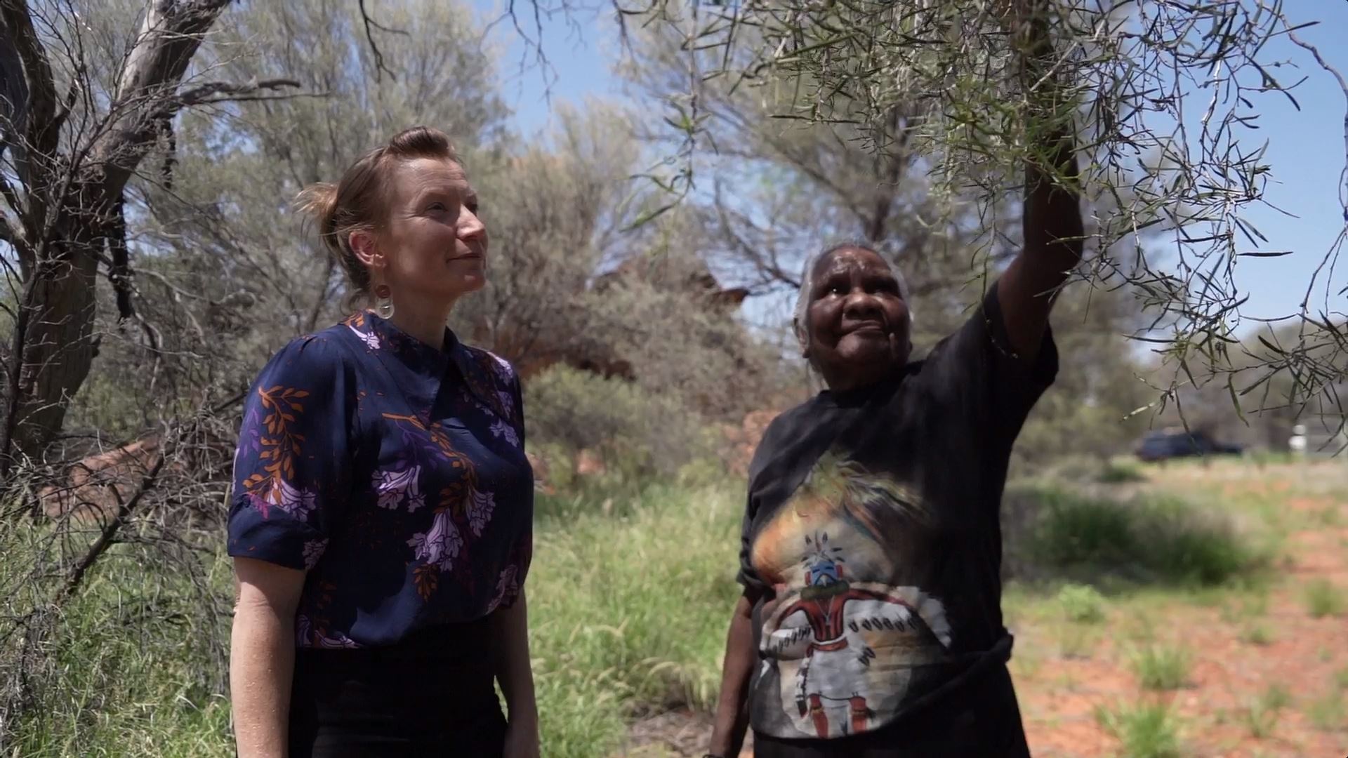 An older Aboriginal woman points up to leaves on a tree as a younger woman watches on.