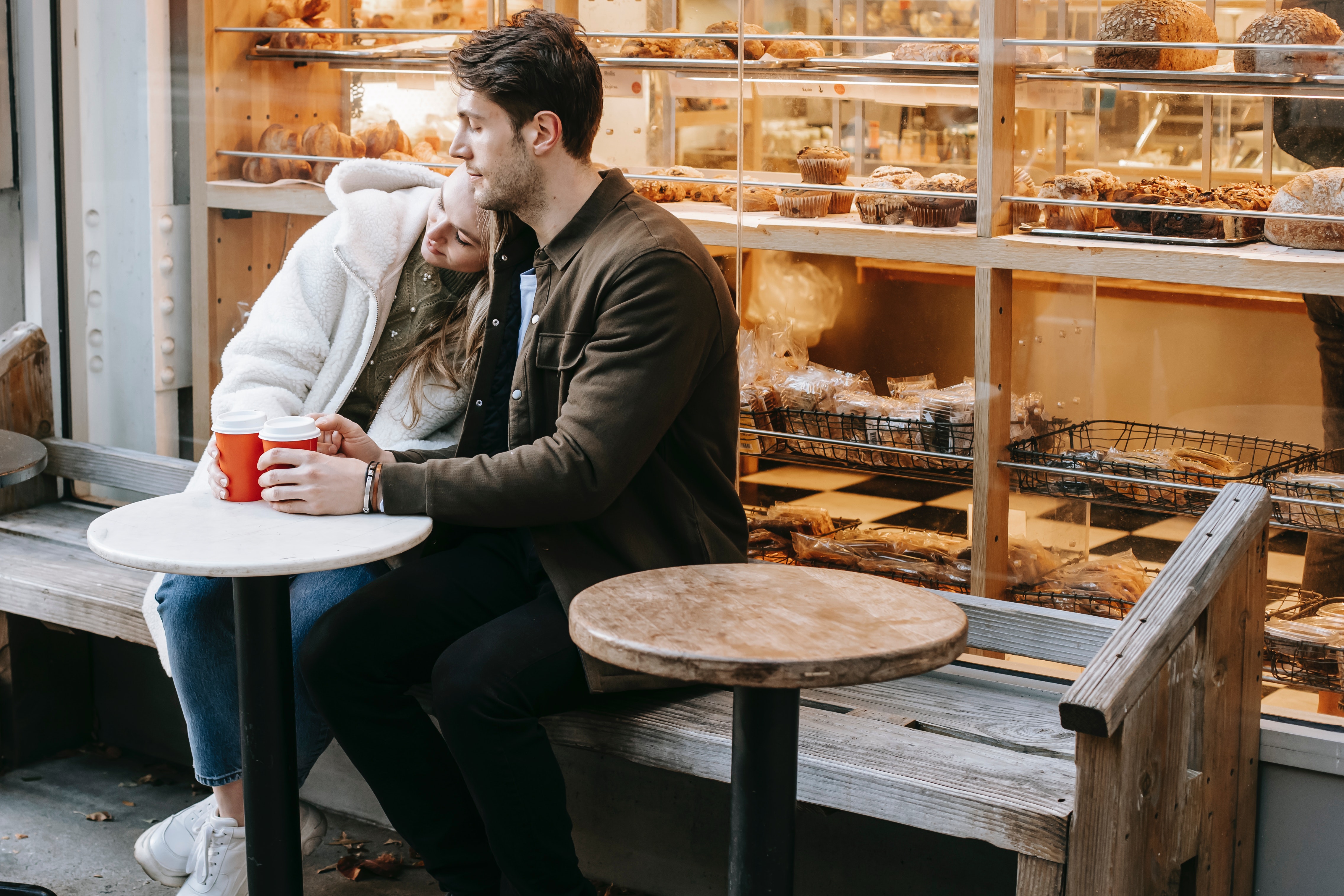 A woman rests her head on a man's shoulder as they have a take-away coffee outside a bakery