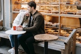 A woman rests her head on a man's shoulder as they have a take-away coffee outside a bakery