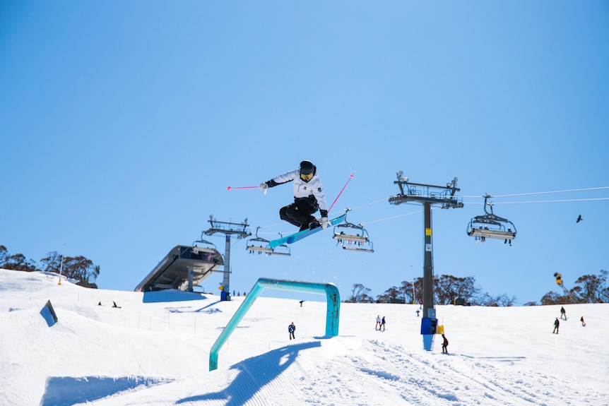 A female skiier in the air with chair lifts in the background.
