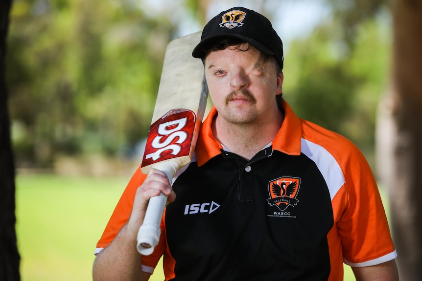 A man in an orange and black polo shirt with a cricket bat over his shoulder.