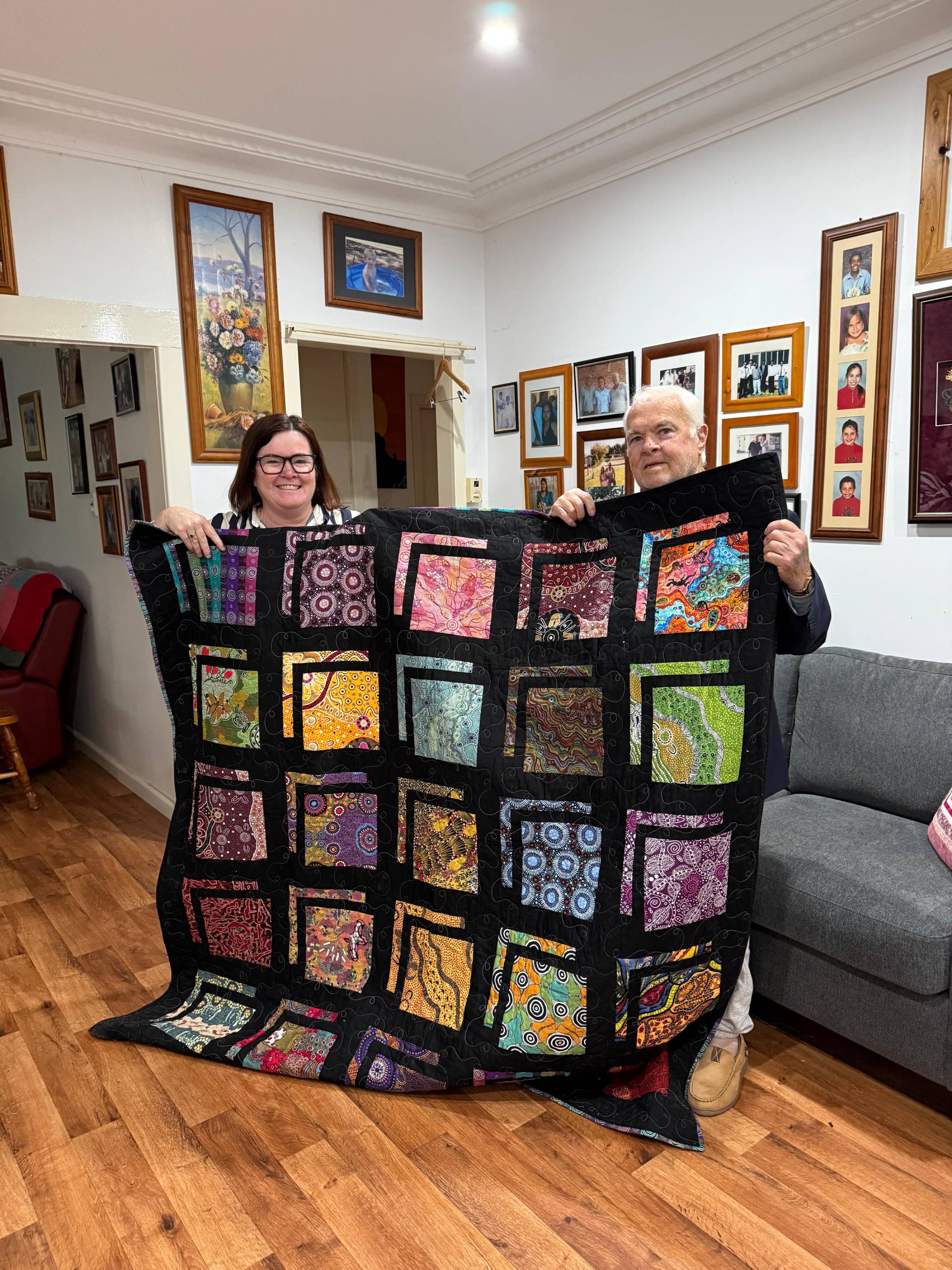 A smiling, dark-haired woman and an older, white-haired man hold up a dark quilt while standing in a living room.