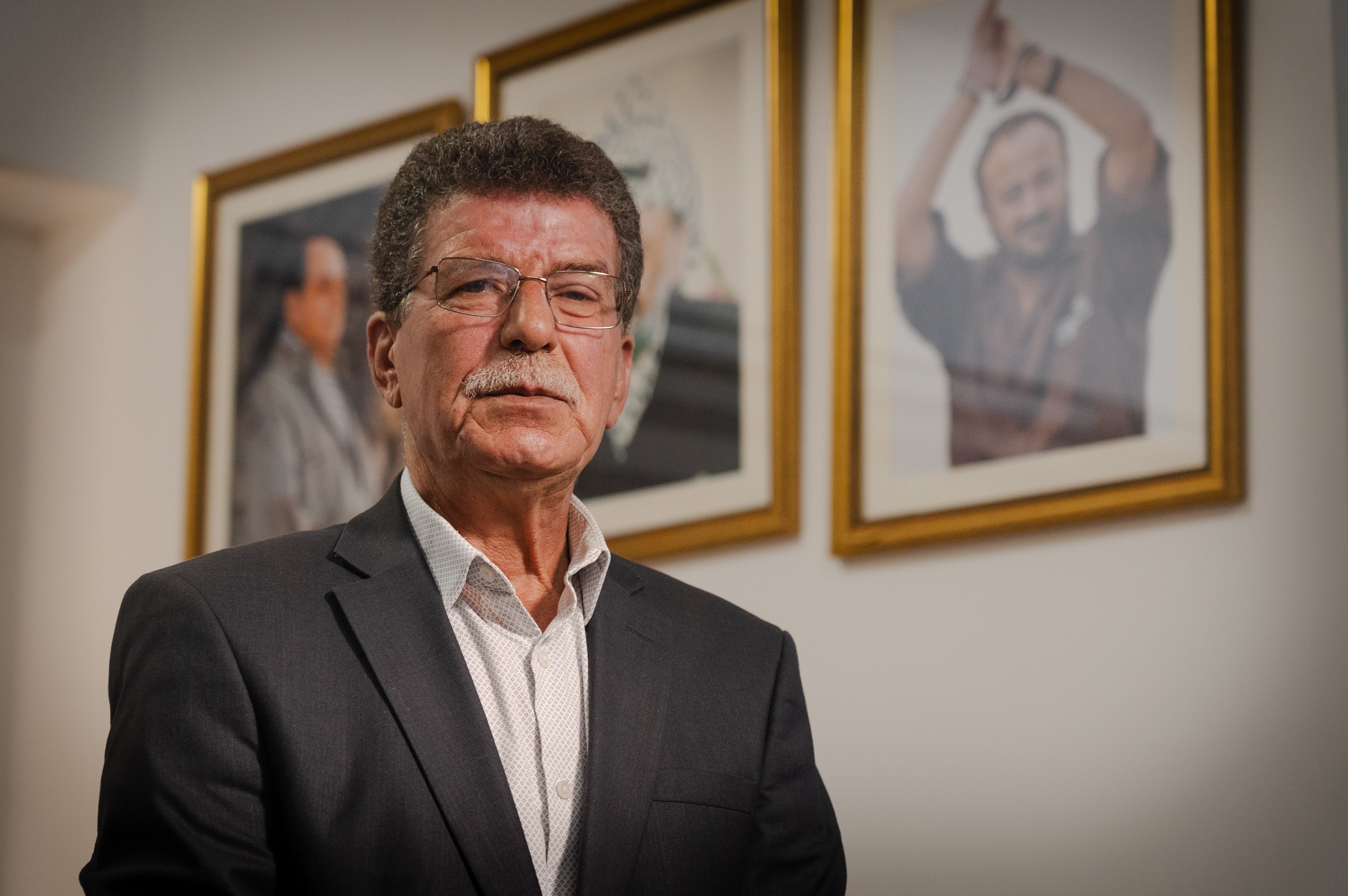 An older man in a suit jacket poses in front of a framed photo of Marwan Barghouti.