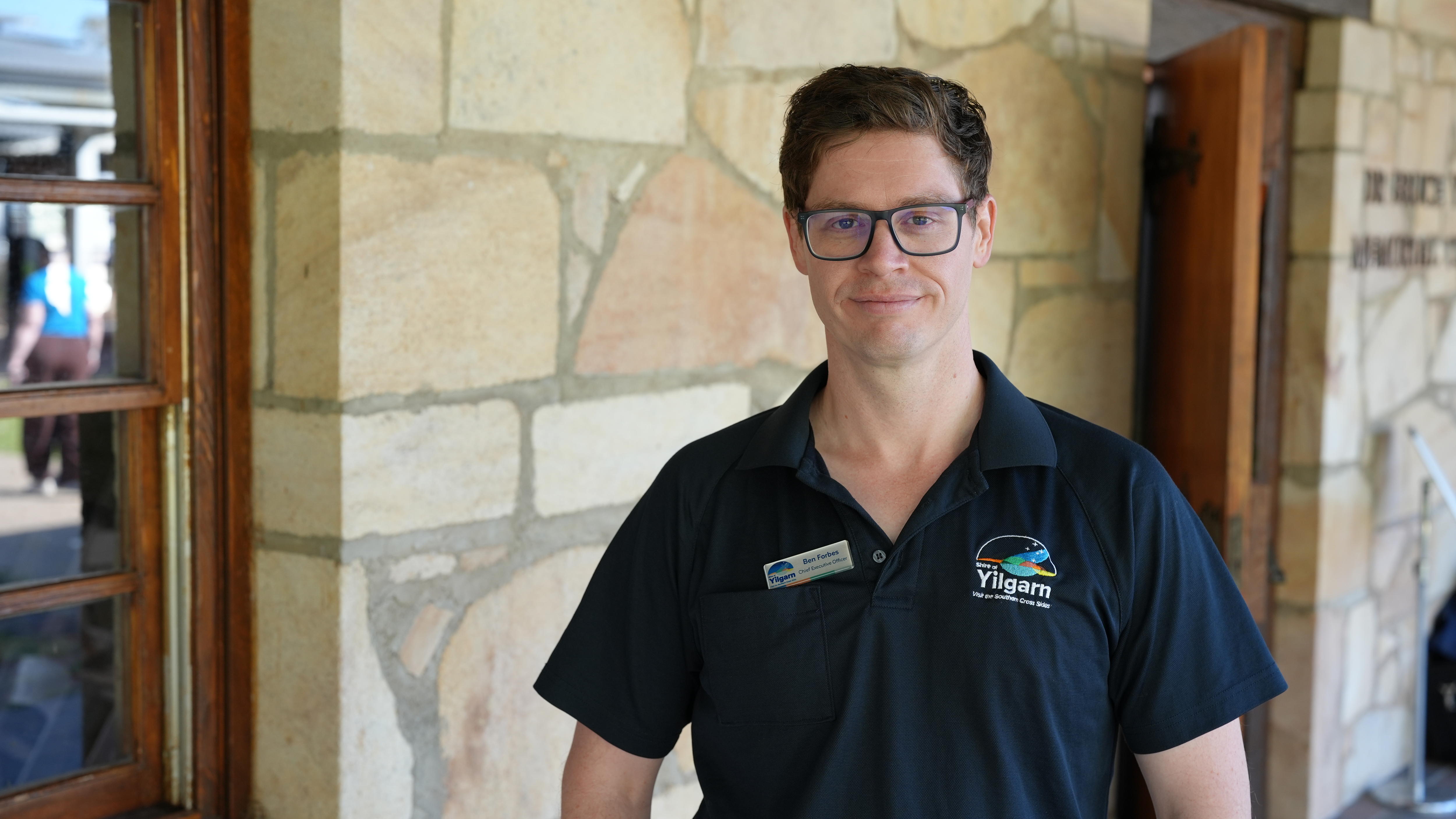 A man (Ben Forbes) wearing a Yilgarn Council polo shirt and glasses, stands in front of a stone wall.