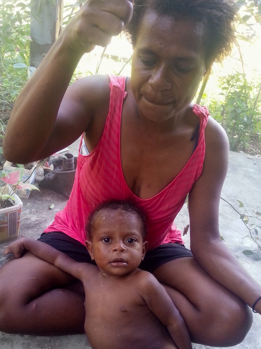 A Papua New Guinean woman in a pink singlet sits cross-legged, with a naked little baby on her lap 