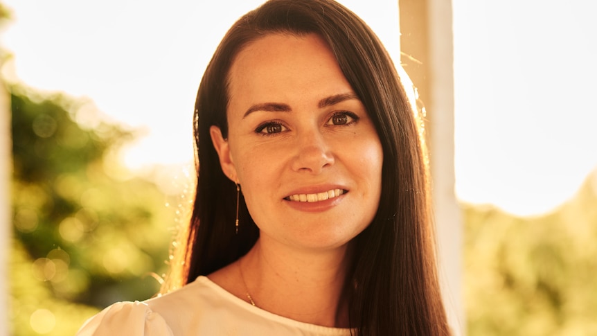 Kylie Moore-Gilbert, with shoulder-length brown hair and white blouse, stands smiling with blurred trees in background.