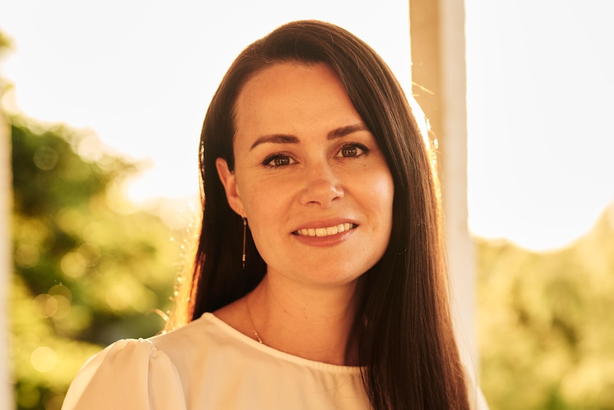 Kylie Moore-Gilbert, with shoulder-length brown hair and white blouse, stands smiling with blurred trees in background.