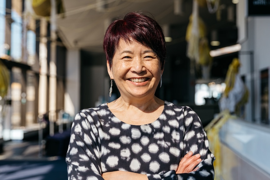 A 60-something Asian Australian woman stands smiling widely in a theatre foyer with her arms folded across her chest