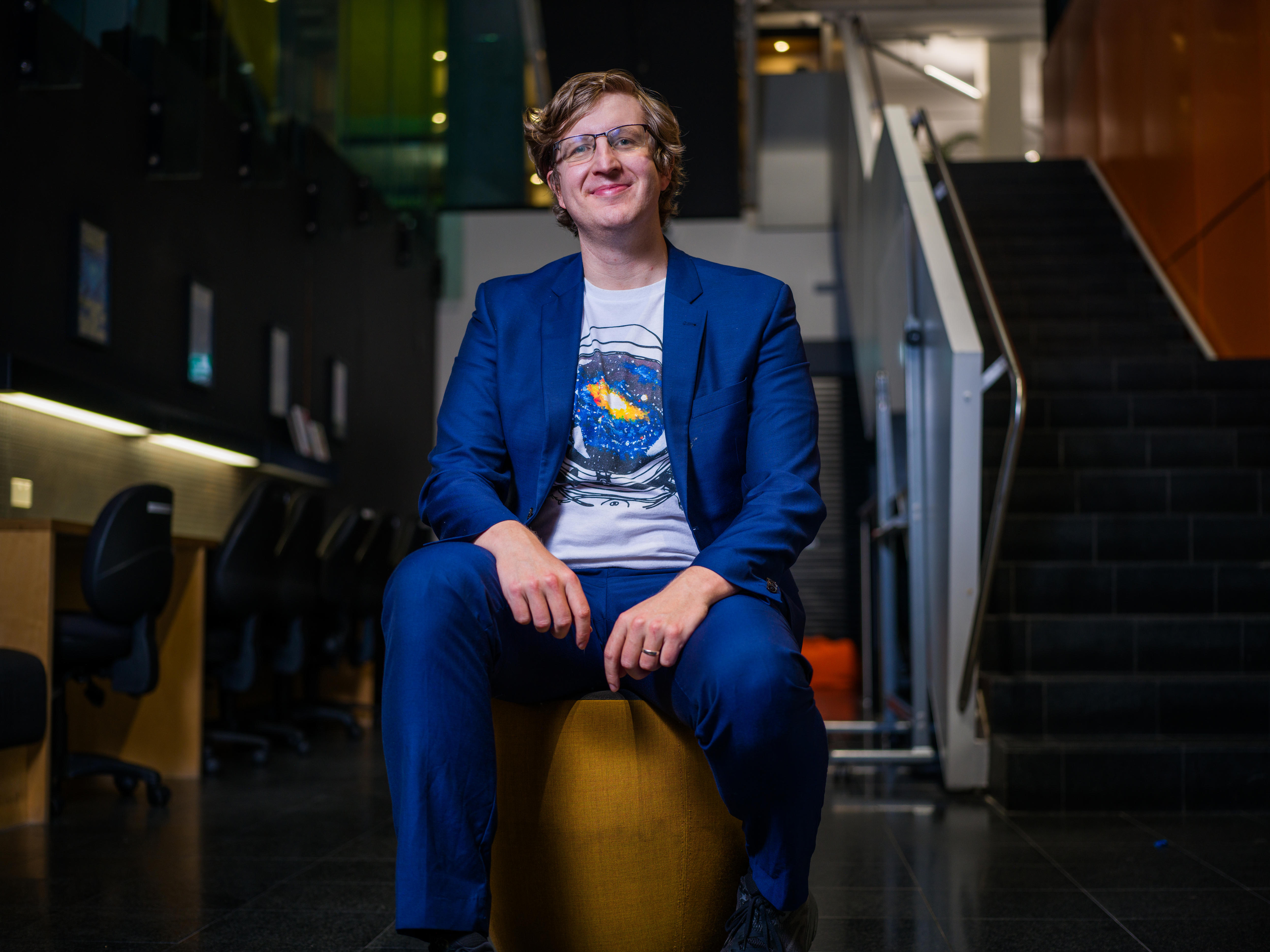 A young man in a blue suit and t-shirt sits on a stool.