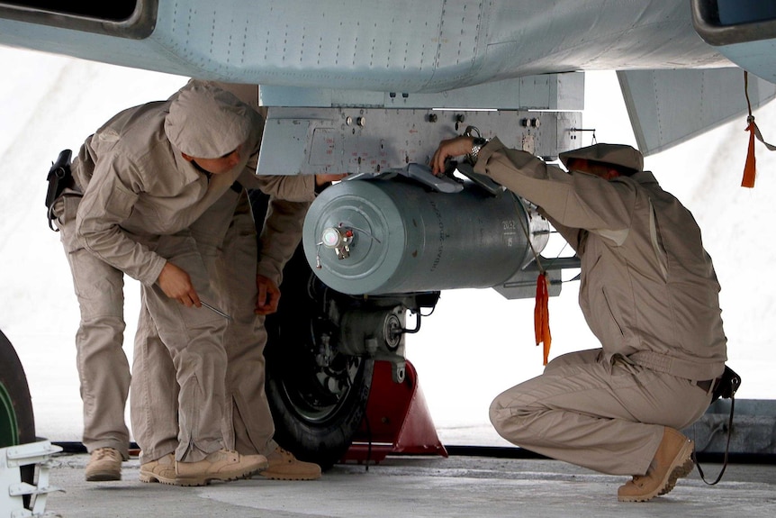 Three ground crew gather around a bomb attached to the underside of a Russian bomber's wing.