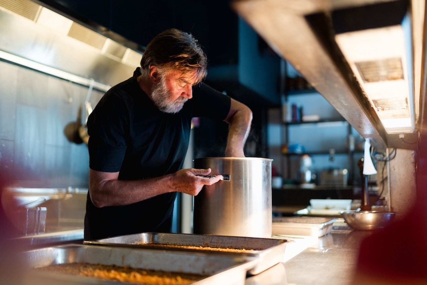 Man stands in commercial kitchen with hands in large pot, surrounded by trays of food