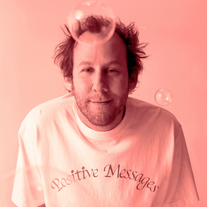 Ben Lee smiles at the camera before a peach background. He wears a white shirt that says 'Positive Messages'. Two bubbles float