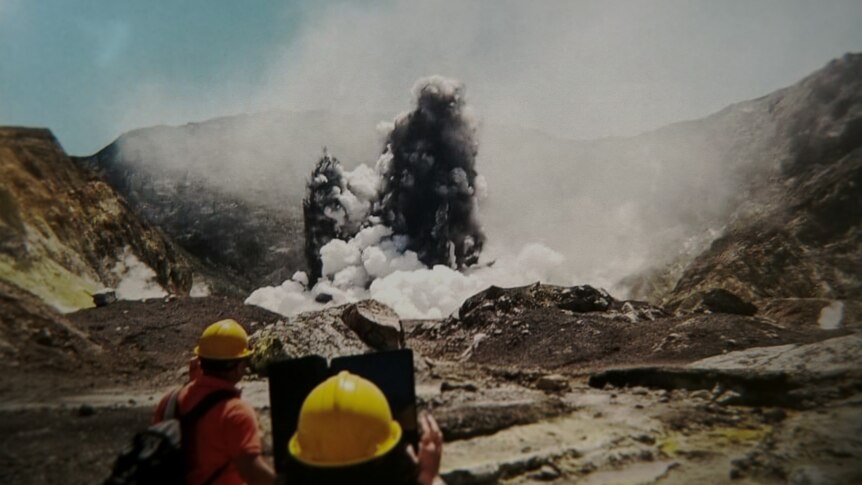 People are seen in the lower part of the image wearing hard hats while the volcano erupts in the background. 
