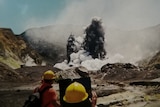 People are seen in the lower part of the image wearing hard hats while the volcano erupts in the background. 