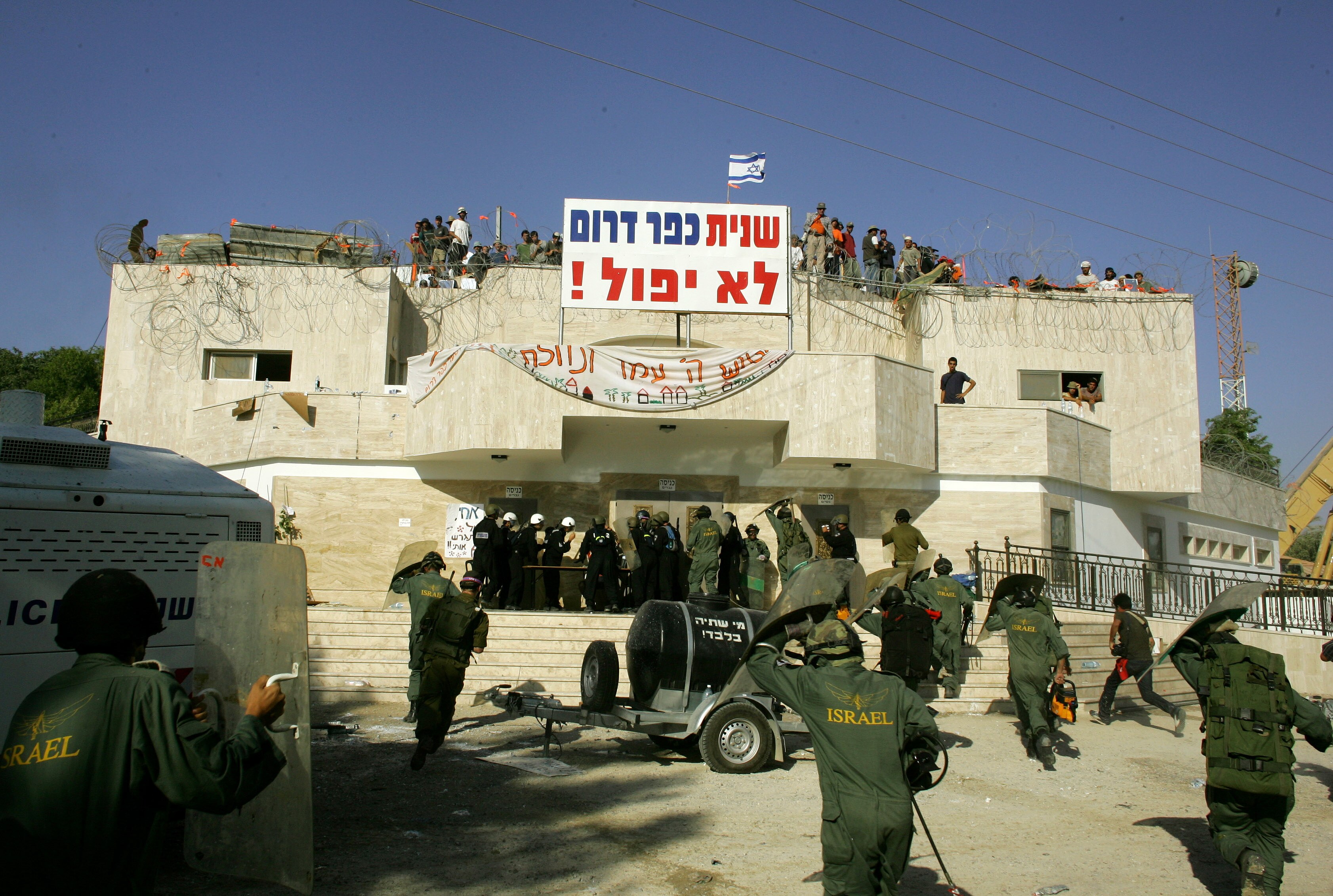 Israeli soldiers wearing green jumpsuits running into a large concrete building topped with barbed wire and protest banners