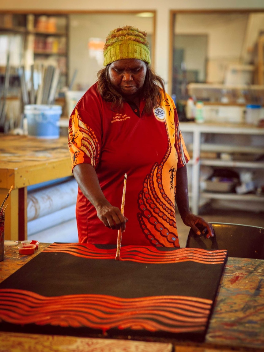 Isobel Major Nampitjinpa paints a canvas with orange paint. She is wearing a red polo and a yellow beanie.