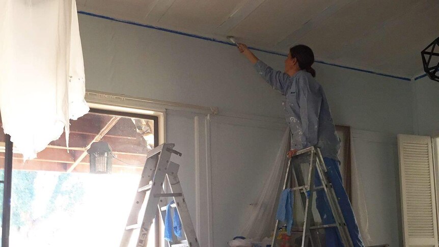 A woman in a paint-splattered shirt on a ladder painting the ceiling