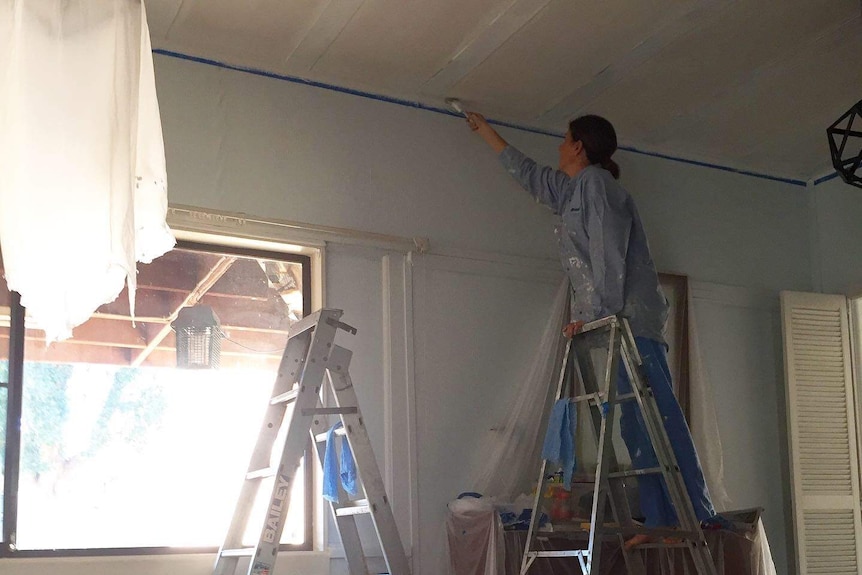 A woman in a paint-splattered shirt on a ladder painting the ceiling