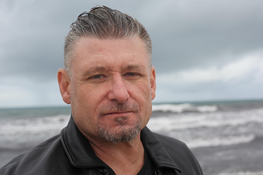 A close up of a man with the ocean and a grey sky behind him. 