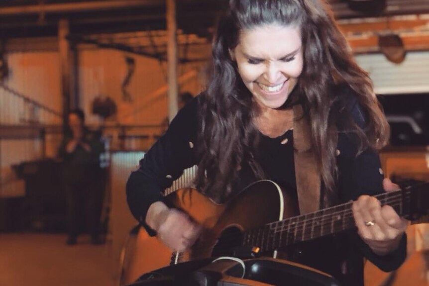 A female guitarist with dark hair smiles as she plays guitar