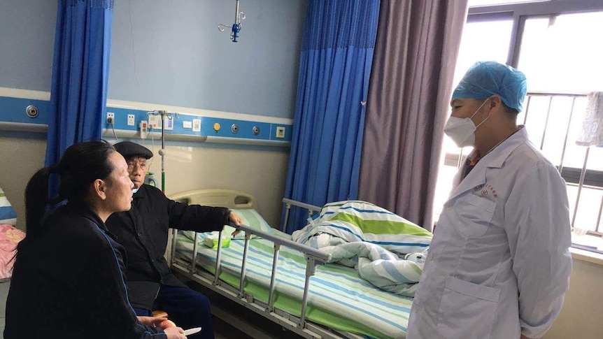 A doctor wearing a lab coat, cap and a mask speaks to patients in their hospital room.