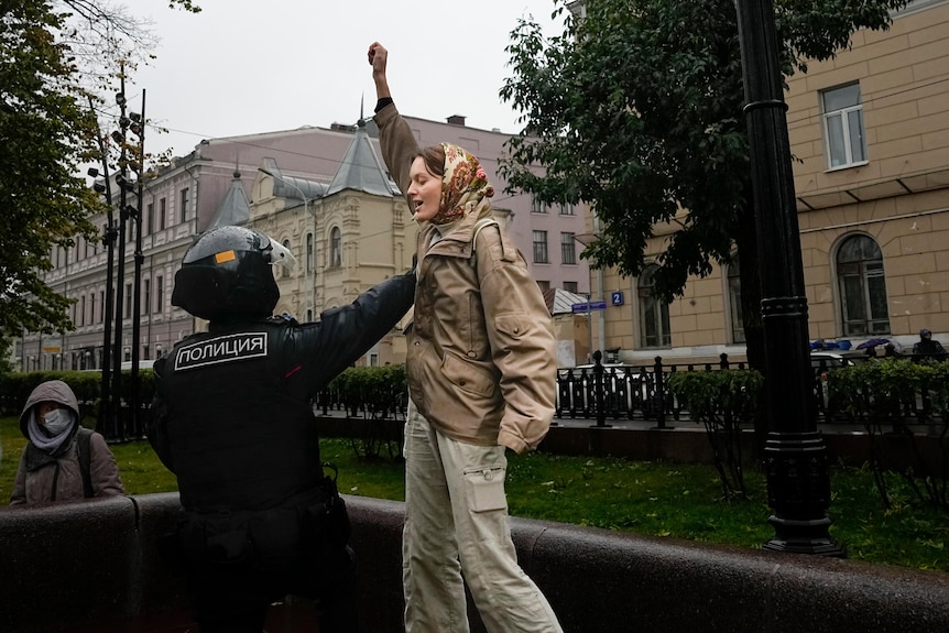 A young white woman in brown clothing raises a fist as she's grasped by a black-clad police officer.