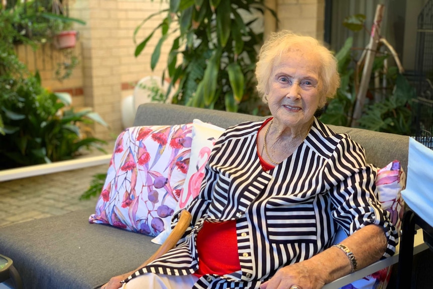 An elderly woman in a bright coloured top and jacket sits smiling in a lounge chair surrounded by colourful cushions and plants.