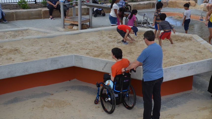 A boy in a wheelchair next to a raised sand pit at a playground