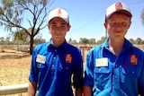 Two boys with blue shirts are standing next to each other. Cattle yards are in the background.