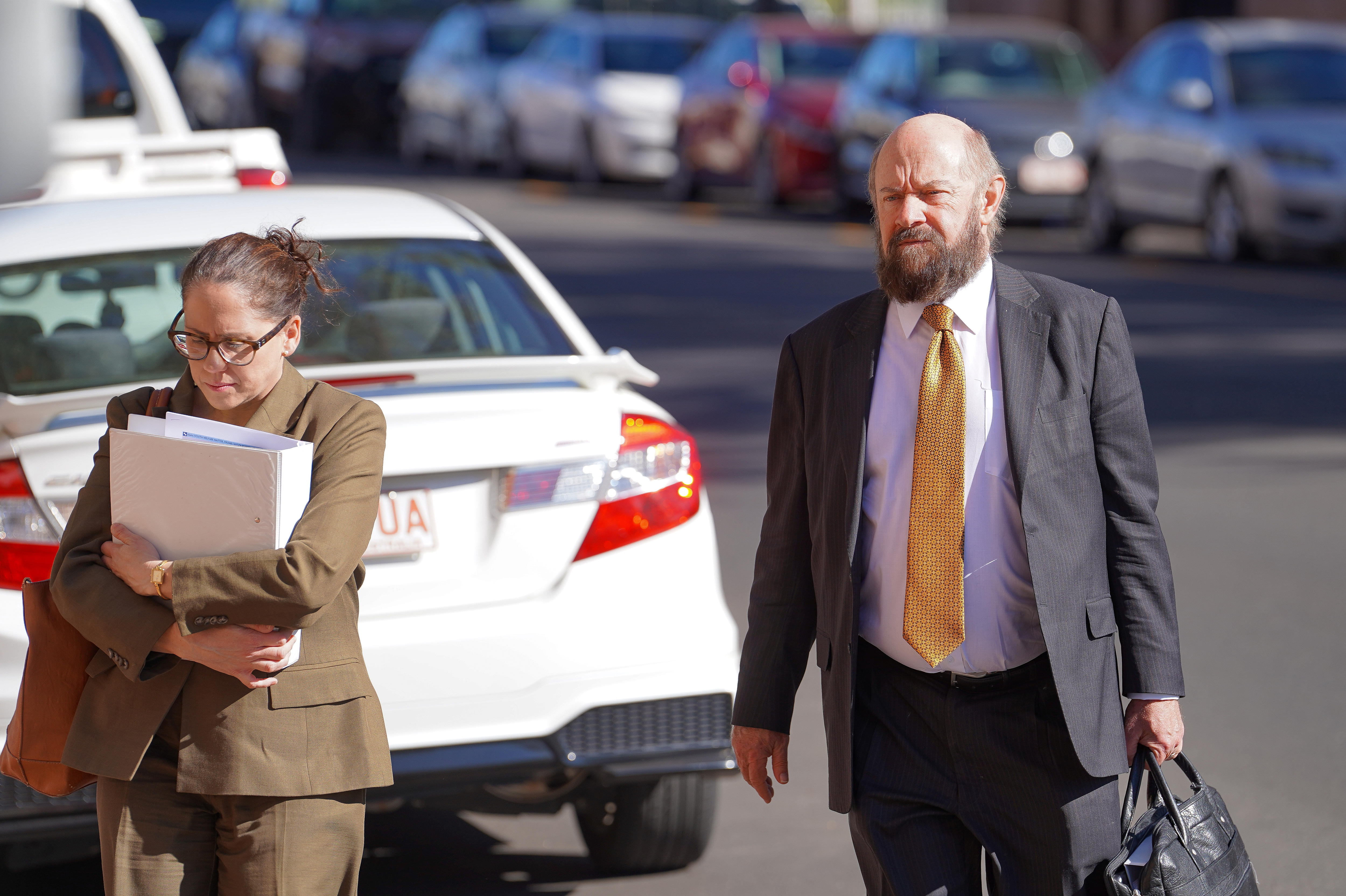 A woman and a man, dressed formally in suits, walking across a road, with rows of cars parked behind them.