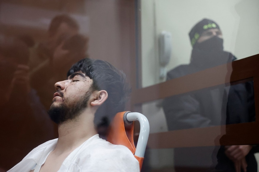 A man with his eyes closed sitting in a chair in a glass box, as a guard with his face covered stands nearby and looks on.