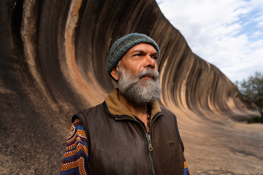 A bearded, older man stands in front of a rock shaped like a wave.