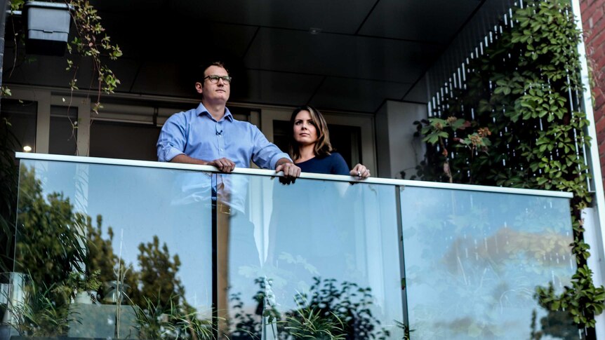 Man in blue shirt and glasses next to woman with blue shirt, standing on apartment balcony with greenery and glass barrier.