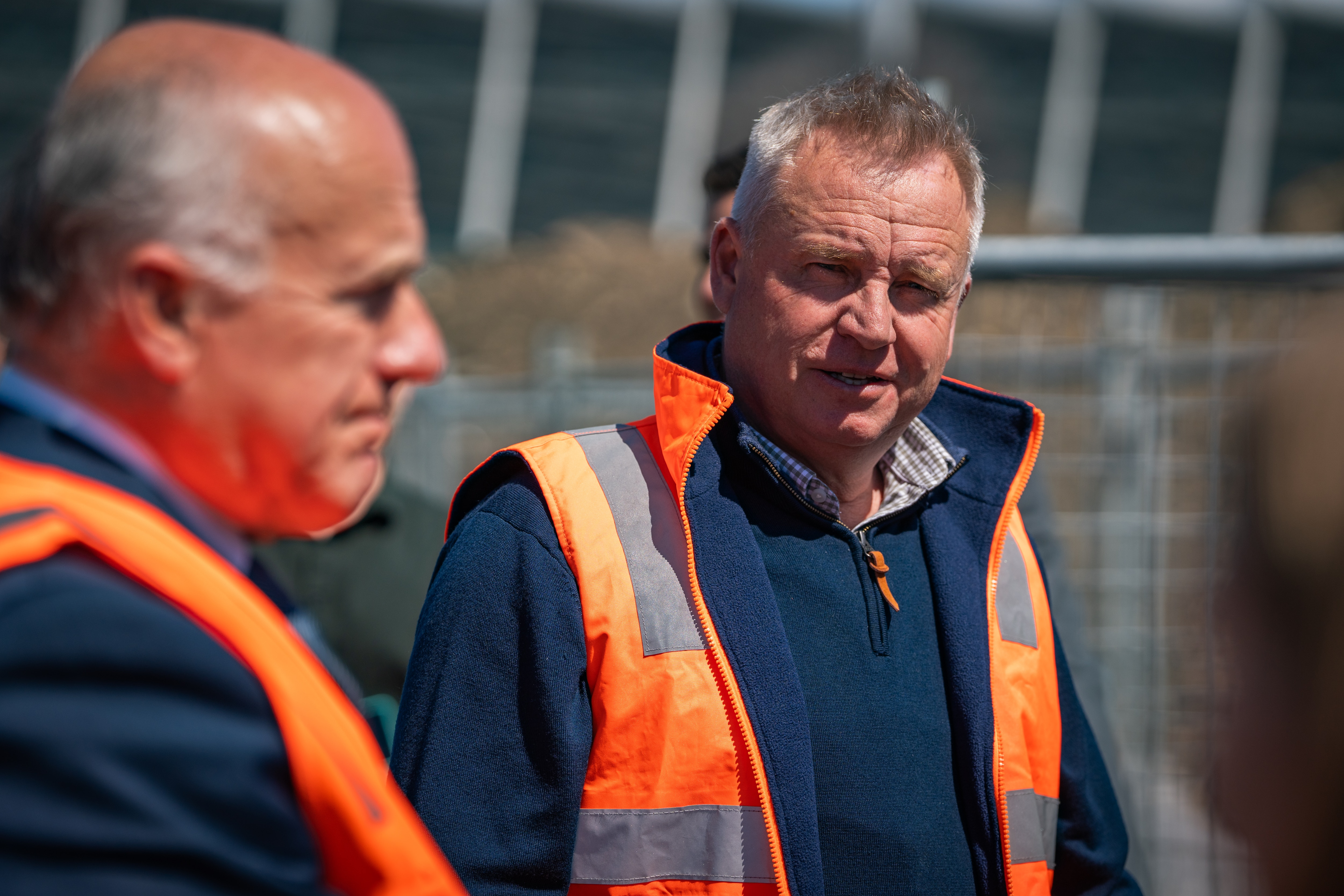 A group of smiling people standing around in high vis in front of an empty dirt lot which is being prepared to become stadium