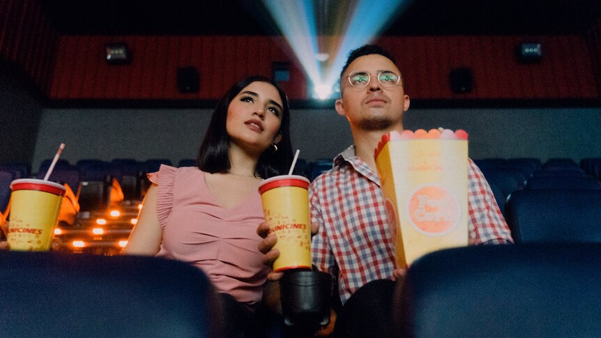 A man and woman sit on plush red seats in a cinema, holding popcorn and drinks.