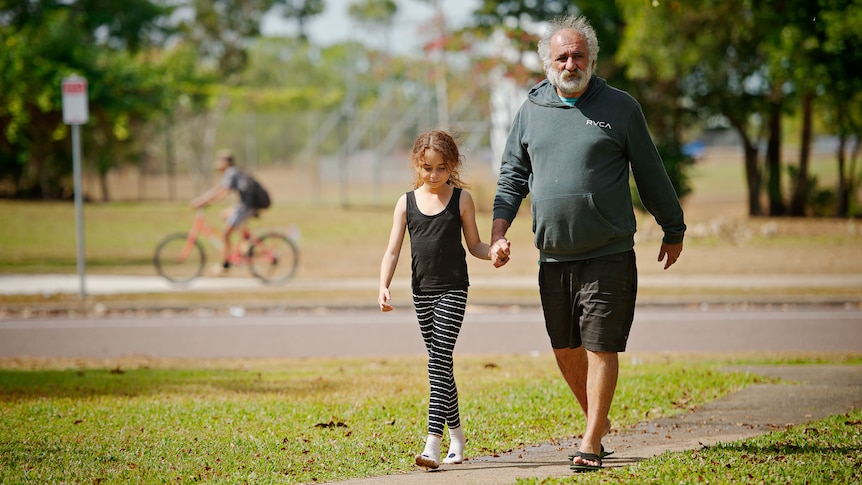 A man wearing a jumper is walking on a footpath with a young girl. They are holding hands as they look to the distance. 