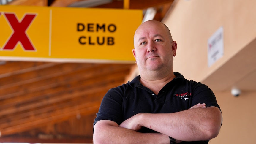 A bald man in black polo shirt stands with his hands crossed in front of a yellow sign reading ' Demo Club'. 