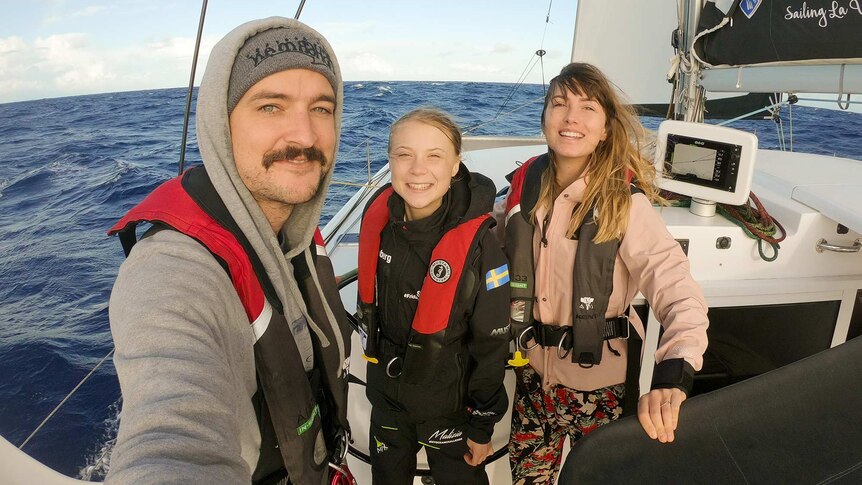 Riley Whitelum, Greta Thunberg and Elayna Carausu pose for a photo standing on the deck of a catamaran at sea.
