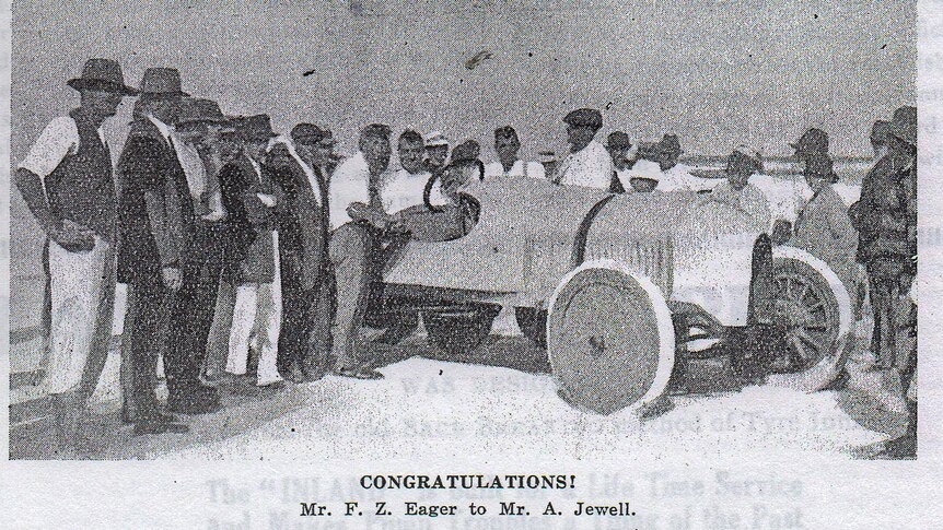 Historic photograph of race car on a beach surrounded by men wearing hats.
